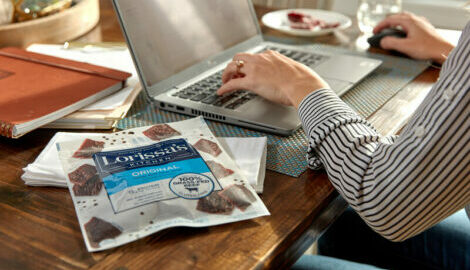 Woman on her laptop working from home with a pack of Lorissa's kitchen Original Steak Strips next to her.