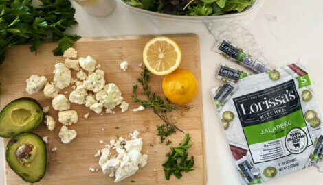 Closeup of a marble dining table and a wooden cutting board. The cutting board has cauliflower, avocado, lemons, and rosemary, and is featured next to a 5-pack of Lorissa's Kitchen Jalapeno Beef Sticks.